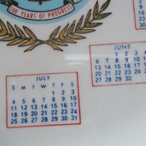 May include: A white ceramic plate with calendar months for June and July. The plate has a blue and gold emblem with the text "200 Years of Progress". The calendar dates are in a red border.