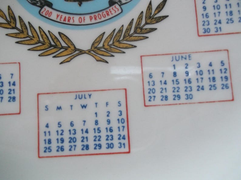 May include: A white ceramic plate with calendar months for June and July. The plate has a blue and gold emblem with the text "200 Years of Progress". The calendar dates are in a red border.