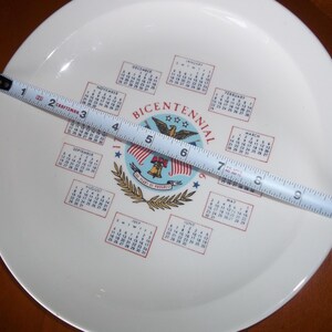 May include: A white plate with a Bicentennial design, featuring a central emblem with an eagle, flags, and a bell. The plate has a diameter of approximately 10 inches, as measured by a tape measure.