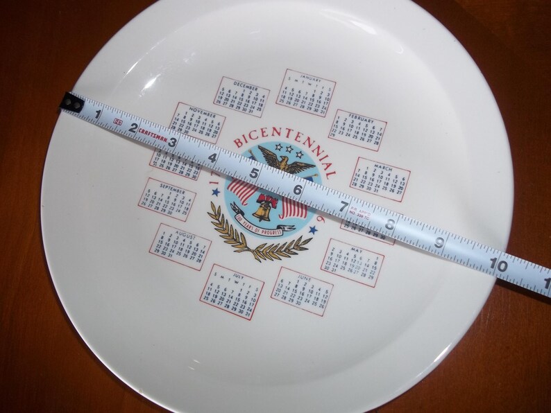 May include: A white plate with a Bicentennial design, featuring a central emblem with an eagle, flags, and a bell. The plate has a diameter of approximately 10 inches, as measured by a tape measure.