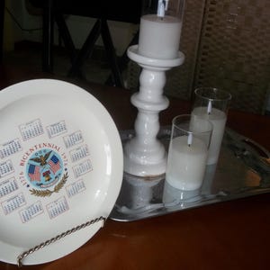 May include: A white plate with a calendar design commemorating the Bicentennial of 1776. A white candle holder with a lit candle, two glass candle holders with lit candles, and a silver tray are also present.
