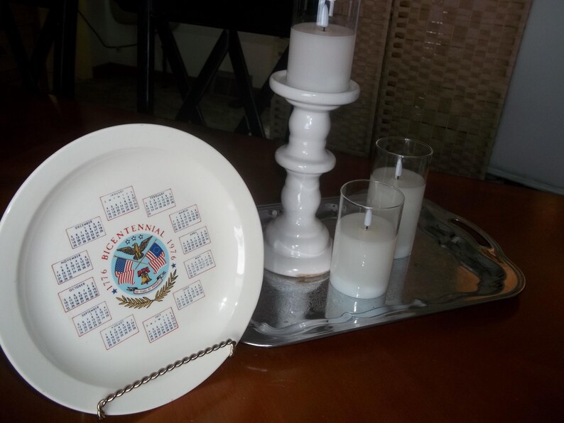 May include: A white plate with a calendar design commemorating the Bicentennial of 1776. A white candle holder with a lit candle, two glass candle holders with lit candles, and a silver tray are also present.