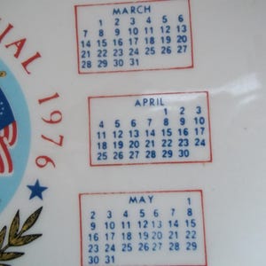 May include: A white ceramic plate with a patriotic design commemorating the 1976 Bicentennial. The plate features the American flag, a blue star, and calendar boxes for March, April, and May.