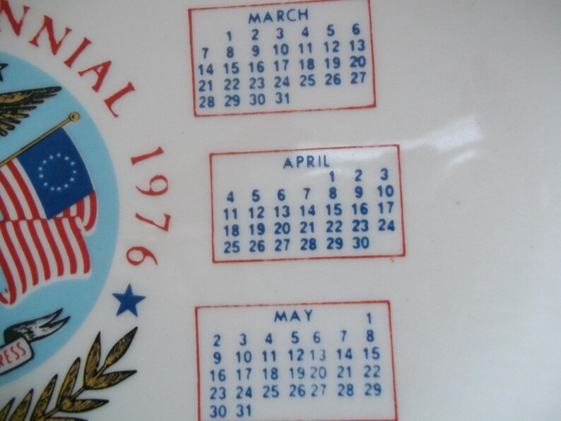 May include: A white ceramic plate with a patriotic design commemorating the 1976 Bicentennial. The plate features the American flag, a blue star, and calendar boxes for March, April, and May.
