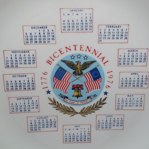 May include: A white ceramic plate featuring a central emblem with the text "BICENTENNIAL 1976". The emblem includes the American and colonial flags, an eagle, and a bell. Calendars for each month are arranged around the emblem.