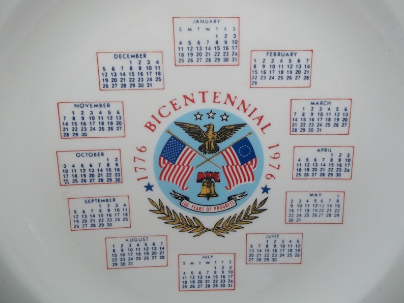 May include: A white ceramic plate featuring a central emblem with the text "BICENTENNIAL 1976". The emblem includes the American and colonial flags, an eagle, and a bell. Calendars for each month are arranged around the emblem.