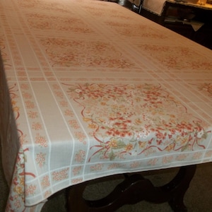 May include: A rectangular tablecloth with a floral pattern in shades of peach, red, and green on a cream background. The design includes floral clusters and ribbon-like accents, creating a vintage aesthetic. The tablecloth is draped over a dark wooden table.