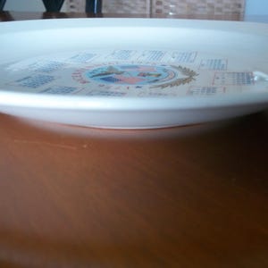 May include: A white ceramic plate with a decorative design. The plate features a central emblem with text and calendar-like elements around the rim. The plate is set on a brown wooden surface.