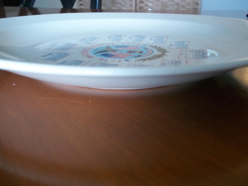 May include: A white ceramic plate with a decorative design. The plate features a central emblem with text and calendar-like elements around the rim. The plate is set on a brown wooden surface.