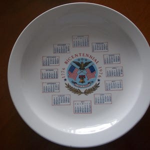 May include: White ceramic plate with a Bicentennial 1776-1976 design. The plate features a central emblem with an eagle, flags, and a bell, surrounded by calendars for each month. A decorative piece.