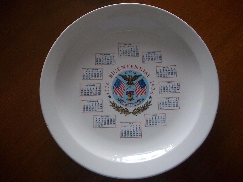 May include: White ceramic plate with a Bicentennial 1776-1976 design. The plate features a central emblem with an eagle, flags, and a bell, surrounded by calendars for each month. A decorative piece.