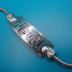 May include: A silver bracelet featuring a rectangular charm with the word "Mom" in cursive. The charm is decorated with a floral motif and is attached to a snake chain. The bracelet has heart-shaped details with small clear stones.