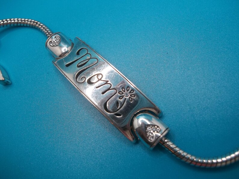 May include: A silver bracelet featuring a rectangular charm with the word "Mom" in cursive. The charm is decorated with a floral motif and is attached to a snake chain. The bracelet has heart-shaped details with small clear stones.