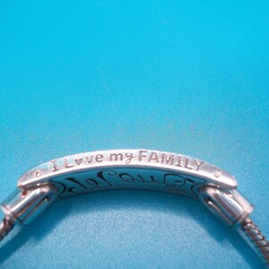 May include: Silver bracelet with the words "I love my FAMILY" inscribed. The bracelet has a flexible, woven chain design. The background is blue.