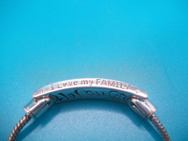 May include: Silver bracelet with the words "I love my FAMILY" inscribed. The bracelet has a flexible, woven chain design. The background is blue.