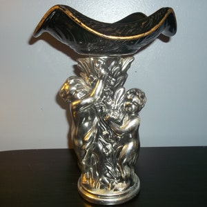 May include: A silver cherub statue with a black and gold bowl on top. The cherubs are holding a floral design.