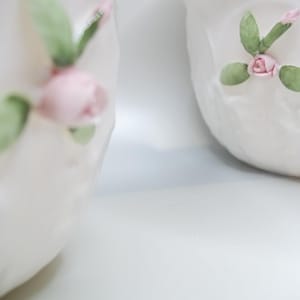 May include: Two white ceramic cups with floral designs. Each cup features a pink rose and green leaves.