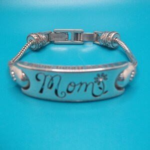 May include: Silver bracelet featuring the word "Mom" in cursive, with a flower detail. The bracelet has a decorative clasp and floral accents. The background is a solid blue.