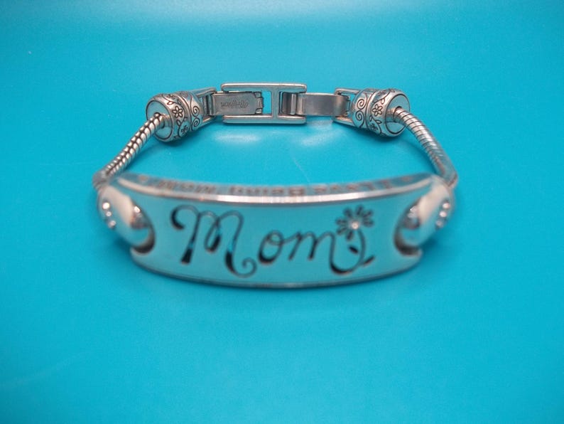 May include: Silver bracelet featuring the word "Mom" in cursive, with a flower detail. The bracelet has a decorative clasp and floral accents. The background is a solid blue.