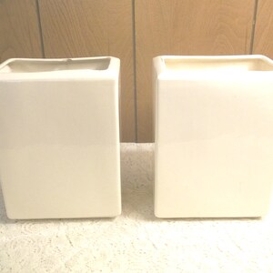 May include: Two white ceramic square planters, side by side, on a white surface.