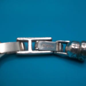 May include: Close-up of a silver bracelet clasp against a blue backdrop. The rectangular clasp has a hinged design, with the brand name visible. The bracelet features a textured, snake-like chain.