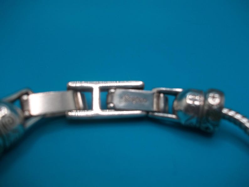 May include: Close-up of a silver bracelet clasp against a blue backdrop. The rectangular clasp has a hinged design, with the brand name visible. The bracelet features a textured, snake-like chain.