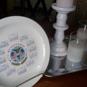 May include: A white plate with a Bicentennial 1776-1976 design, featuring a calendar and American flags. A white candle holder with a pink candle and two glass candles sit on a silver tray.