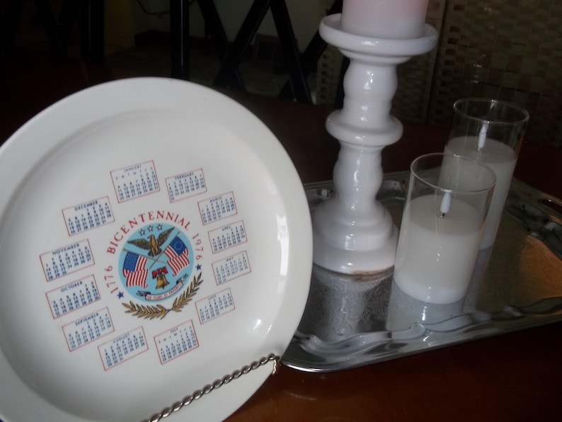 May include: A white plate with a Bicentennial 1776-1976 design, featuring a calendar and American flags. A white candle holder with a pink candle and two glass candles sit on a silver tray.