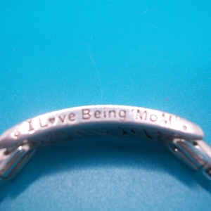 May include: Silver bracelet with the inscription "I Love Being Mom". The bracelet has a snake chain design and is set against a bright blue background. The bracelet is a piece of jewellery.