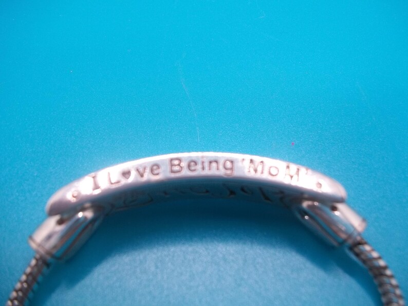 May include: Silver bracelet with the inscription "I Love Being Mom". The bracelet has a snake chain design and is set against a bright blue background. The bracelet is a piece of jewellery.