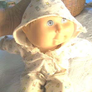 May include: A Cabbage Patch Kid doll wearing a white hooded onesie with a small animal print. The doll has blue eyes and a rosy complexion.