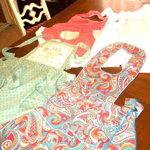 May include: A collection of colorful aprons in various patterns and designs. One apron features a red paisley pattern with blue and orange accents. Another is light green with small floral prints. A third apron is red with white polka dots.