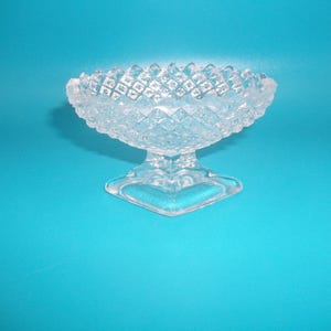 May include: A clear glass pedestal bowl featuring a textured diamond pattern across the exterior surface. The bowl has a scalloped rim and sits on a square base, showcasing a reflective, transparent design with intricate geometric detailing.