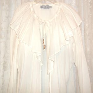 May include: A white cotton blouse with a ruffled neckline and long sleeves. The blouse has a tie at the neck and wooden beads hanging from the tie.