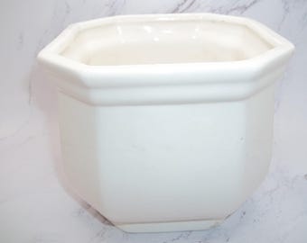 Puede incluir: Una maceta de cerámica blanca sólida con forma octogonal y un borde ancho y acampanado. El recipiente presenta líneas suaves y limpias con un acabado brillante, proporcionando un contenedor simple y funcional para plantas con un interior profundo.