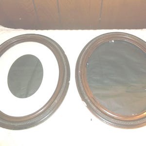 May include: Two oval-shaped picture frames with ornate wood trim. The frame on the left has a white mat and a smaller oval opening. The frame on the right has a dark glass insert.