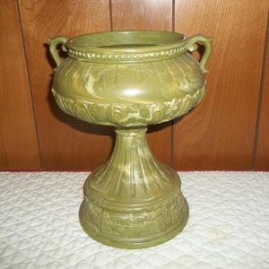 May include: A green decorative urn with two handles, a wide bowl, and a pedestal base. The urn has a detailed, textured design with a fluted base and ornate carvings. The color is a muted olive green.