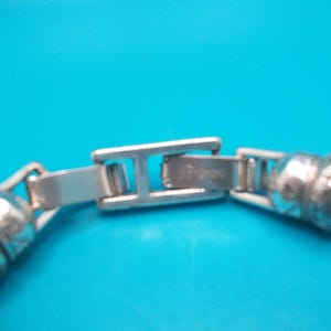 May include: Close-up of a silver bracelet clasp against a bright blue background. The clasp is rectangular with a hinged design, connecting to a textured, rounded chain. The bracelet appears to be made of metal.