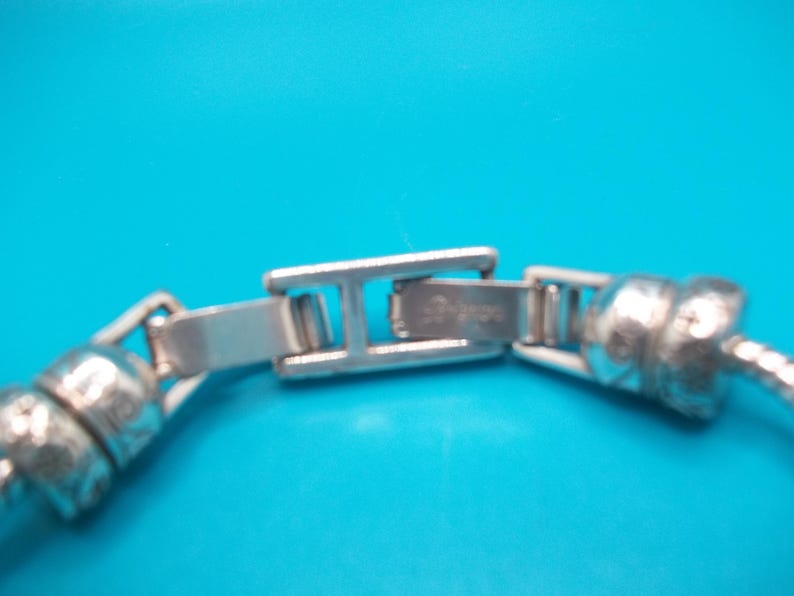May include: Close-up of a silver bracelet clasp against a bright blue background. The clasp is rectangular with a hinged design, connecting to a textured, rounded chain. The bracelet appears to be made of metal.