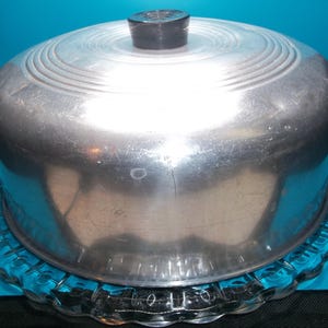 May include: A vintage aluminum cake carrier with a black knob on top, resting on a clear, scalloped glass plate. The cake carrier has a domed lid with concentric rings. The background is a solid teal color.