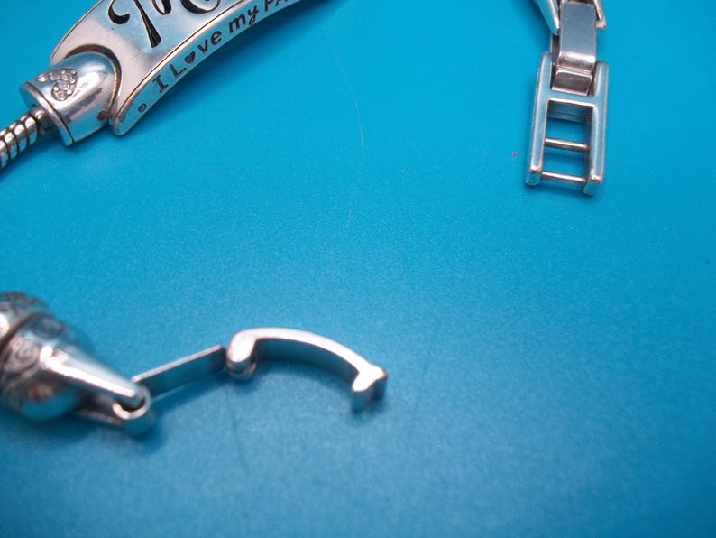 May include: Silver bracelet with a clasp and a decorative charm. The charm has the inscription "I love my..." and a small heart. The bracelet is on a blue background.