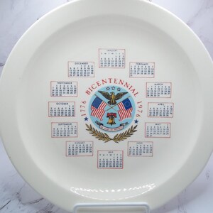 May include: A white ceramic plate with a Bicentennial design. The plate features the years 1776 and 1976, along with the words "Bicentennial" and a central emblem with flags, an eagle, and a bell. Calendars for each month are also displayed.