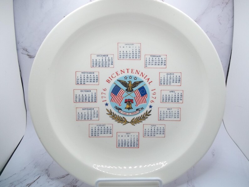 May include: A white ceramic plate with a Bicentennial design. The plate features the years 1776 and 1976, along with the words "Bicentennial" and a central emblem with flags, an eagle, and a bell. Calendars for each month are also displayed.