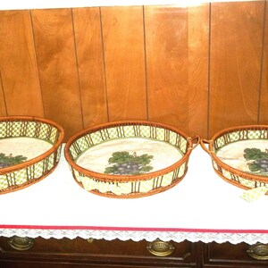 May include: Three oval wicker serving trays with a grape design. The trays have a brown wicker frame, a green and white checkered border, and a white interior with a grape cluster illustration. The trays are on a white surface.