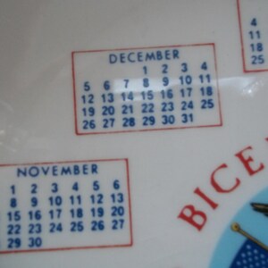 May include: White ceramic plate with calendar months of November and December. The calendar dates are in blue, with red borders. The word "BICENT" is visible in red.