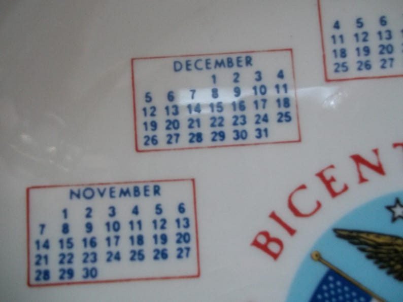 May include: White ceramic plate with calendar months of November and December. The calendar dates are in blue, with red borders. The word "BICENT" is visible in red.