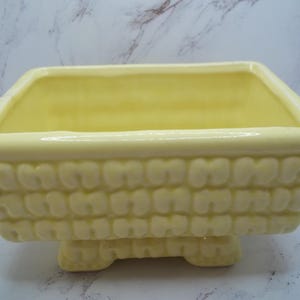 May include: A yellow ceramic planter with a textured, woven design and four short legs. The planter is empty and ready for a plant.