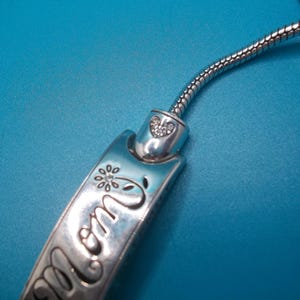 May include: A silver bracelet charm with the word "mom" engraved on it, along with a flower and heart design. The charm is connected to a flexible, silver chain. The background is a solid blue.