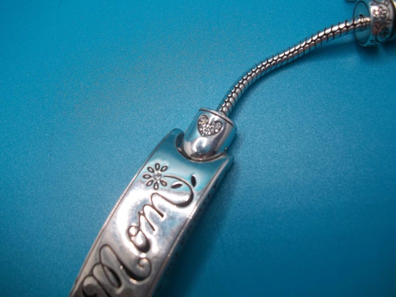 May include: A silver bracelet charm with the word "mom" engraved on it, along with a flower and heart design. The charm is connected to a flexible, silver chain. The background is a solid blue.