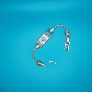 May include: A silver bracelet featuring the word "Mom" engraved on a rectangular charm. The bracelet has a flexible, coiled design with decorative beads and a clasp. The background is a solid teal colour.